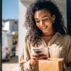woman with curly brown hair receives package and stands at the door of her house with her smartphone in her hand looking at the package tracker app