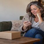 Woman on a sofa checking her phone beside a small parcel