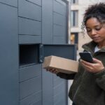 Woman at outdoor parcel lockers holding a parcel and checking her phone