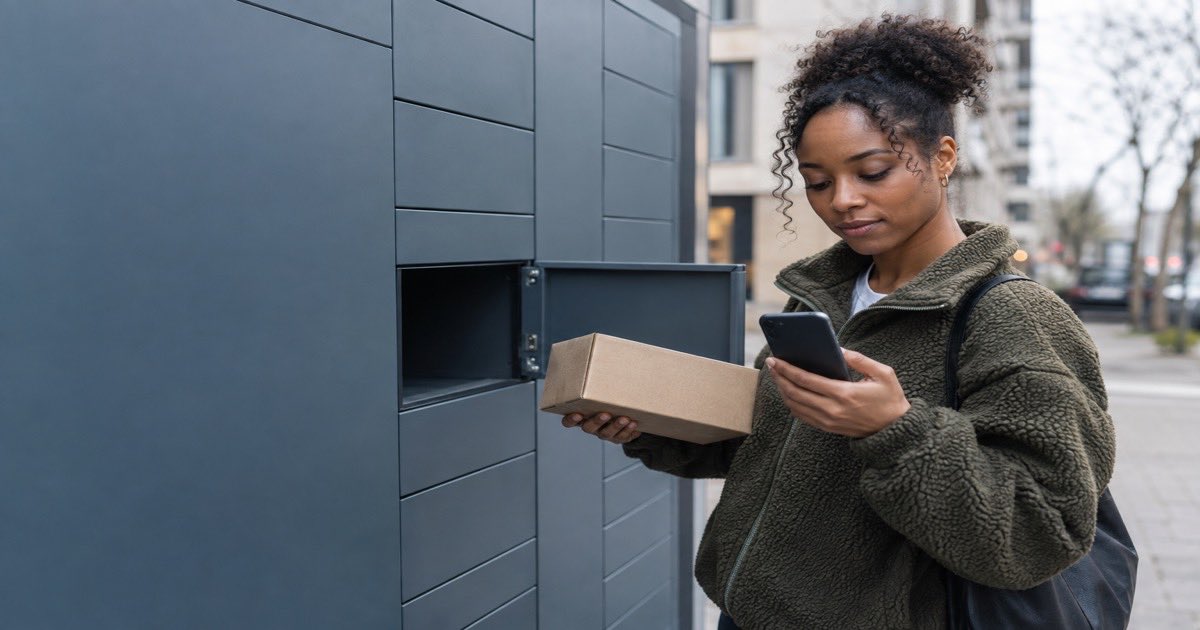 Woman at outdoor parcel lockers holding a parcel and checking her phone