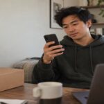 Young man at a home desk checking his phone beside a plain parcel