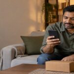 Man on a sofa checking his phone beside a small parcel in the evening
