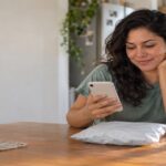 Woman at a kitchen table checking her phone beside a poly mailer