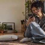 Young person in a bedroom with a clothing rack checking their phone beside a mailer