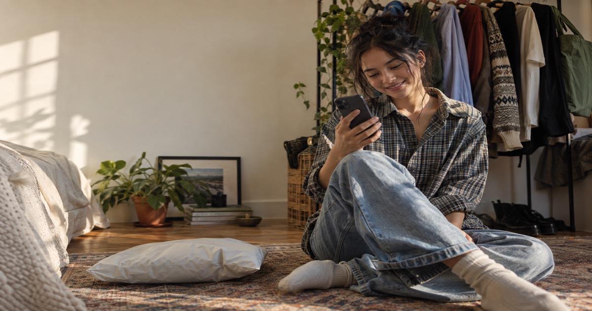 Young person in a bedroom with a clothing rack checking their phone beside a mailer