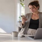 Woman at a kitchen island with a laptop checking her phone while waiting for a parcel